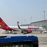 A SpiceJet Boeing 737-8AS grounded on Tarmac with cargo staff handling luggage. An airport building and a clear blue sky in the background.