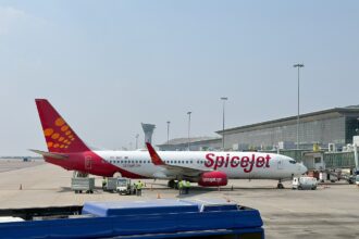 A SpiceJet Boeing 737-8AS grounded on Tarmac with cargo staff handling luggage. An airport building and a clear blue sky in the background.