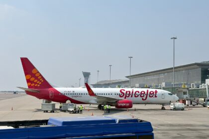 A SpiceJet Boeing 737-8AS grounded on Tarmac with cargo staff handling luggage. An airport building and a clear blue sky in the background.