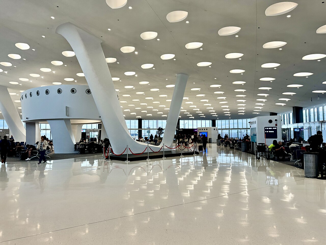 This photo depicts Terminal 5 Concourse in Chicago O'Hare International Airport. The ceiling, floor and architecture is decorated white. The ceiling holds warm calming light emitted from dotted holes lined up in neat rows. The terminal doesn't appear busy and people are only lightly scattered around.