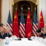 Meeting between China and the US. Chinese and US flags posed against each other in the background. The classic red with yellow stars and blue with white, red stripes and stars stand out against the white table in front with frustrated looking men.