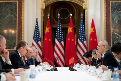 Meeting between China and the US. Chinese and US flags posed against each other in the background. The classic red with yellow stars and blue with white, red stripes and stars stand out against the white table in front with frustrated looking men.