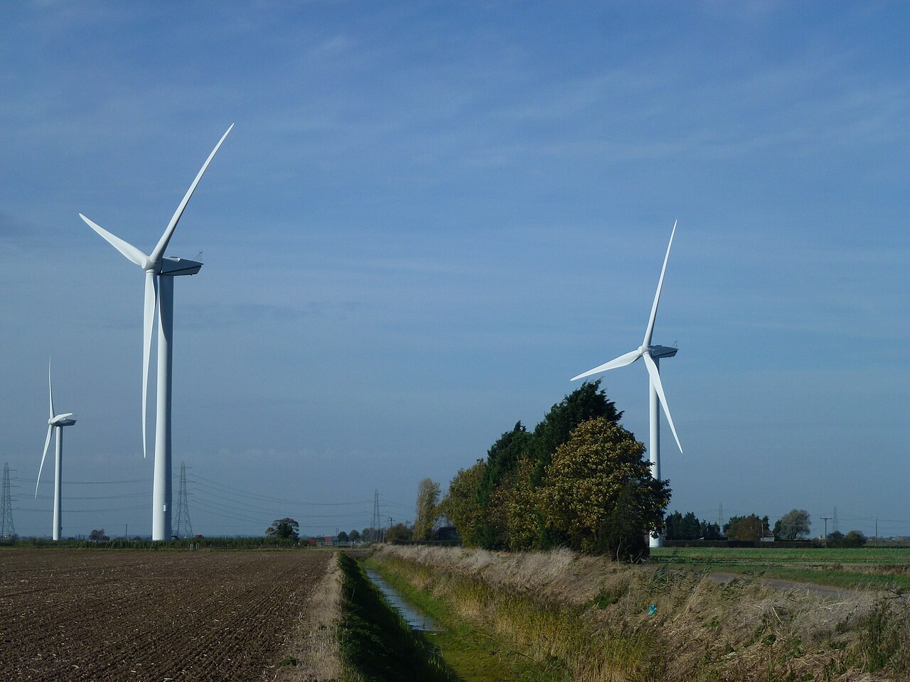 Picture of three white wind turbines in Bicker Fen, Boston Lincolnshire, rotating in two brown and greenish fields. There are a few overgrown shrubs surrounding the fields. The sky is blue and a little bit cloudy.