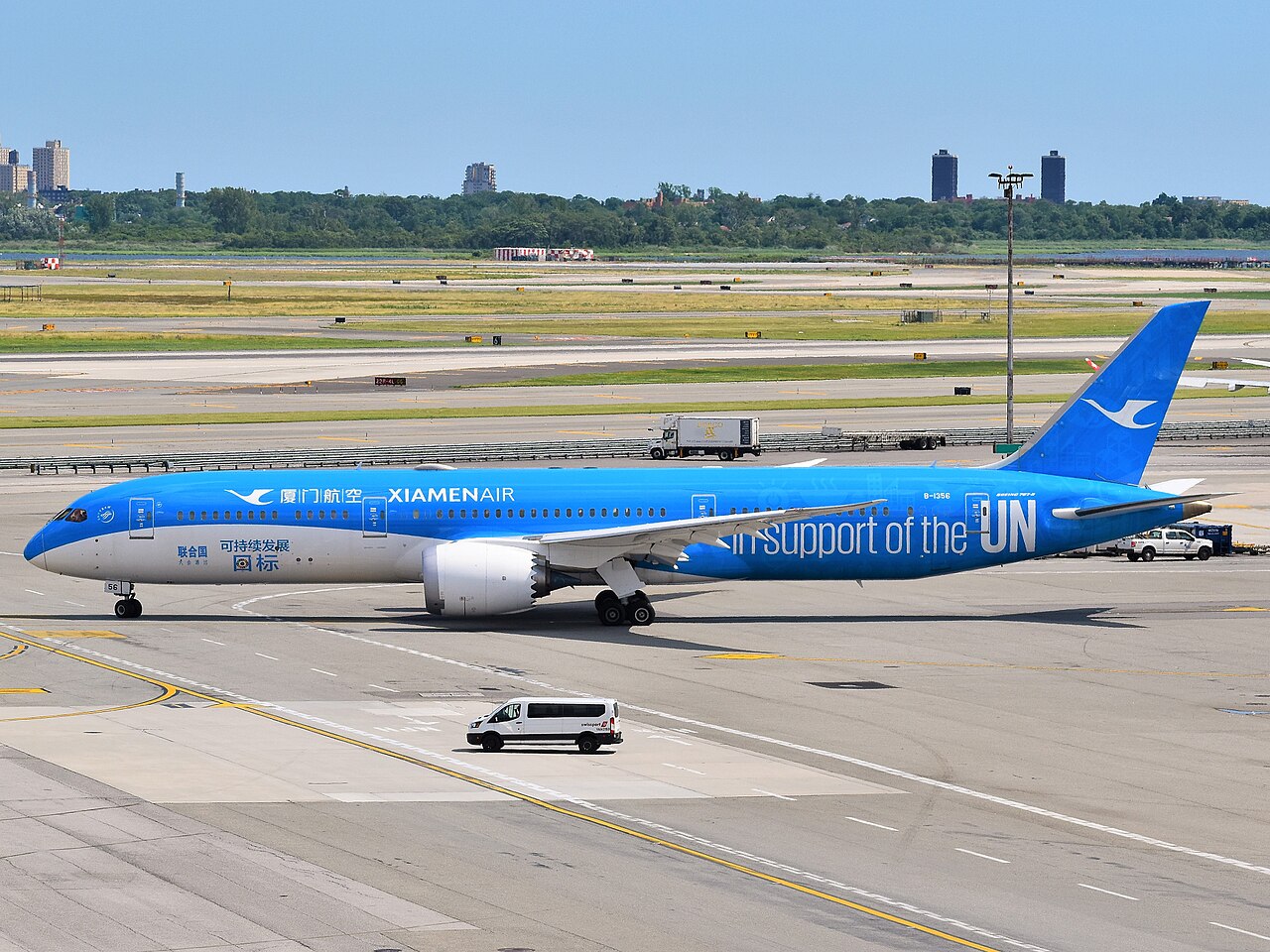 A Chinese enterprise, XiamenAir Boeing 787-9 aircraft, painted in blue and white inscribed with text saying "in support of the UN". The airplane is taxiing out for departure as Flight MF850 to Fuzhou. In the background is a calm bright blue sky and the rest of a large airport run way.