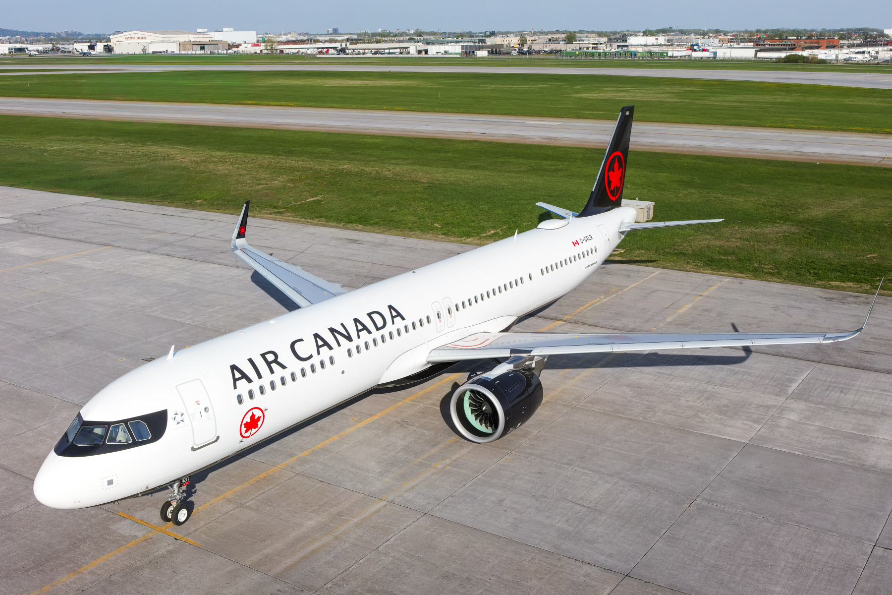 Air Canada branded Airbus A321-XLR parked sideways on the runway with Air Canada maple leaf on tail and body. The sky is blue.