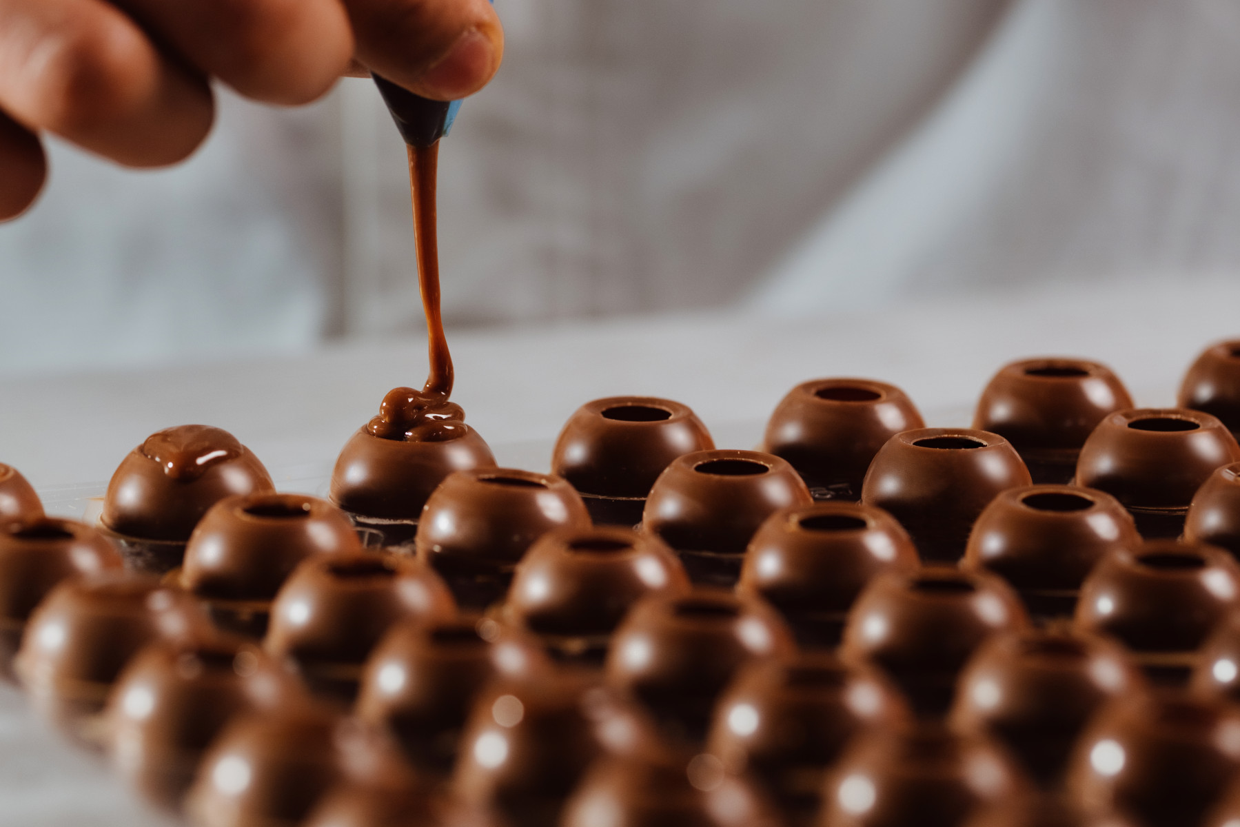 A chocolatier is filling truffles with expert precision, there are many small truffles lined up to be filled.