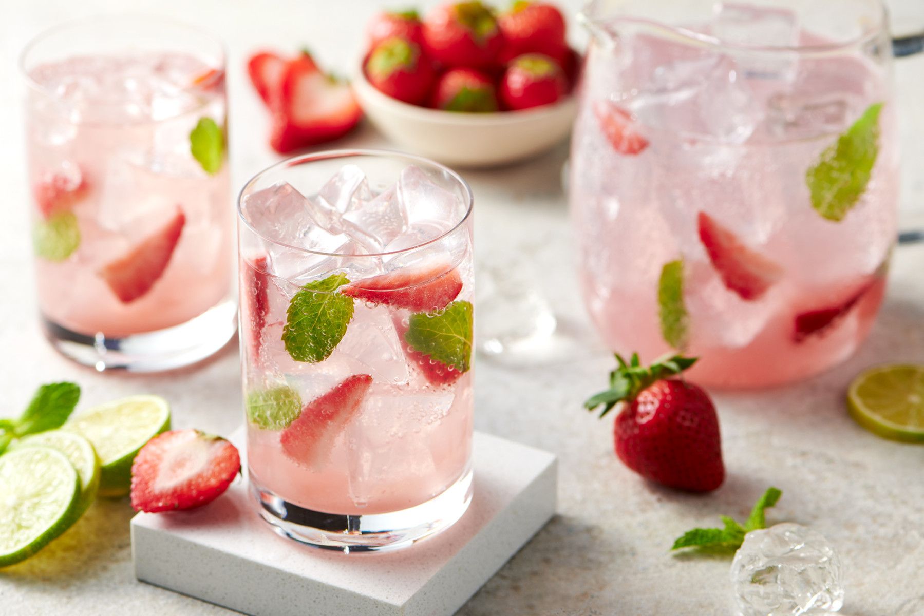 strawberry inspired drinks with ice, mint, lime and more strawberries arranged on a table. the drinks have a slight pink tinge to them.