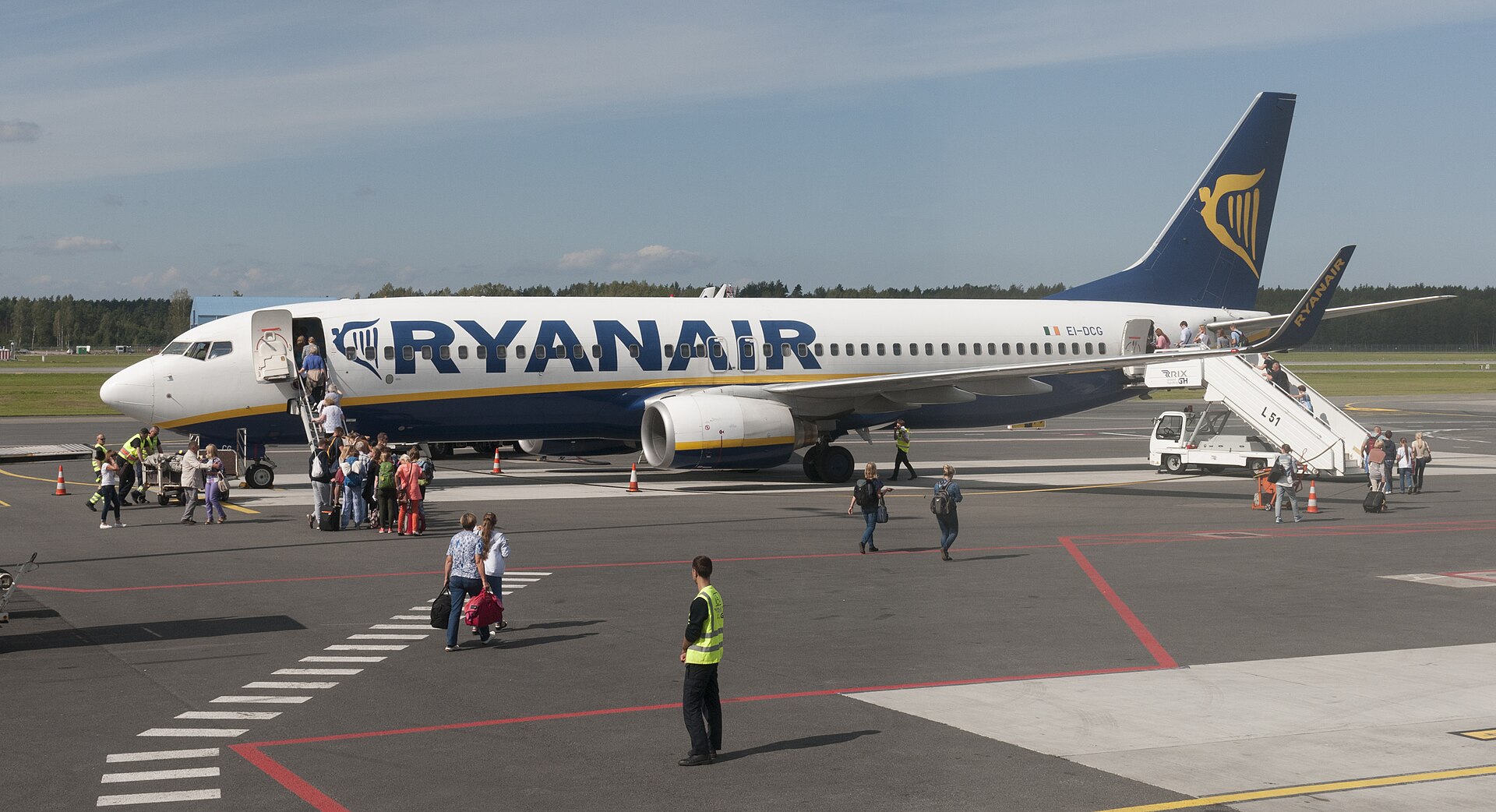 A Ryanair jet is grounded at an airport, and people with suitcases approach and board the flight from stairs ascending from the ground to the front doors. The nose of the aircraft is facing left, the tail on the right. The aircraft is mostly white with a navy blue underbelly, navy blue branding across the fuselage reading 'RYANAIR' and a navy blue tail with the yellow-gold logo on it. The aircraft is in the centre of the image. The foreground is occupied by tarmac ground with lines, as well as people walking across the tarmac to the plane. The background is occupied by a few trees and grass beneath a vast pale blue sky. 