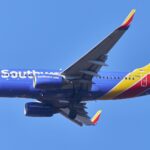 A Southwest Airlines plane in flight. The aircraft is cobalt blue, with white branding along the side of the fuselage. The tail of the aircraft, which is to the right of the image, is bright red and egg-yolk yellow. The background is bright cloudless blue sky.