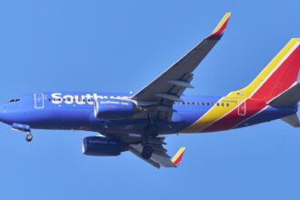 A Southwest Airlines plane in flight. The aircraft is cobalt blue, with white branding along the side of the fuselage. The tail of the aircraft, which is to the right of the image, is bright red and egg-yolk yellow. The background is bright cloudless blue sky.