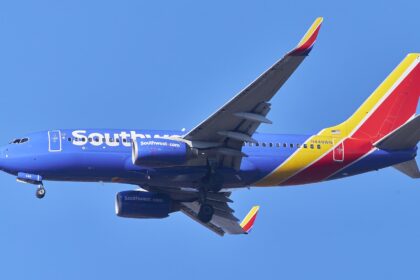 A Southwest Airlines plane in flight. The aircraft is cobalt blue, with white branding along the side of the fuselage. The tail of the aircraft, which is to the right of the image, is bright red and egg-yolk yellow. The background is bright cloudless blue sky.