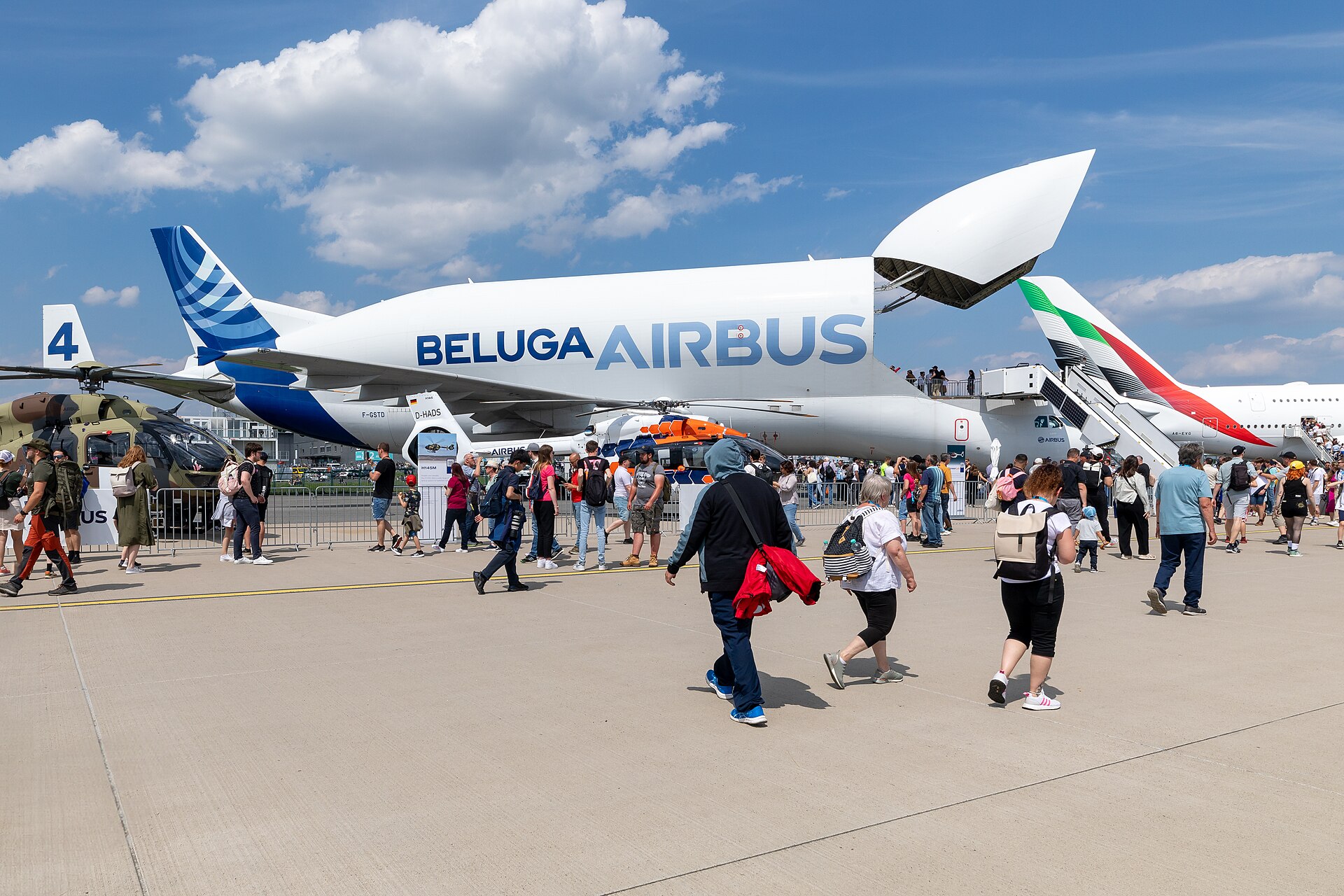 An Airbus Beluga aircraft is grounded and on display at what looks like an aviation fair. The aircraft is in the centre of the image, and is shown from the side a short distance away. The beluga aircraft is open at the front. While mostly white, the aircraft does have blue branding on the tail and along the side. People pass by the aircraft in varying directions. The background shows a blue sky with some clouds.