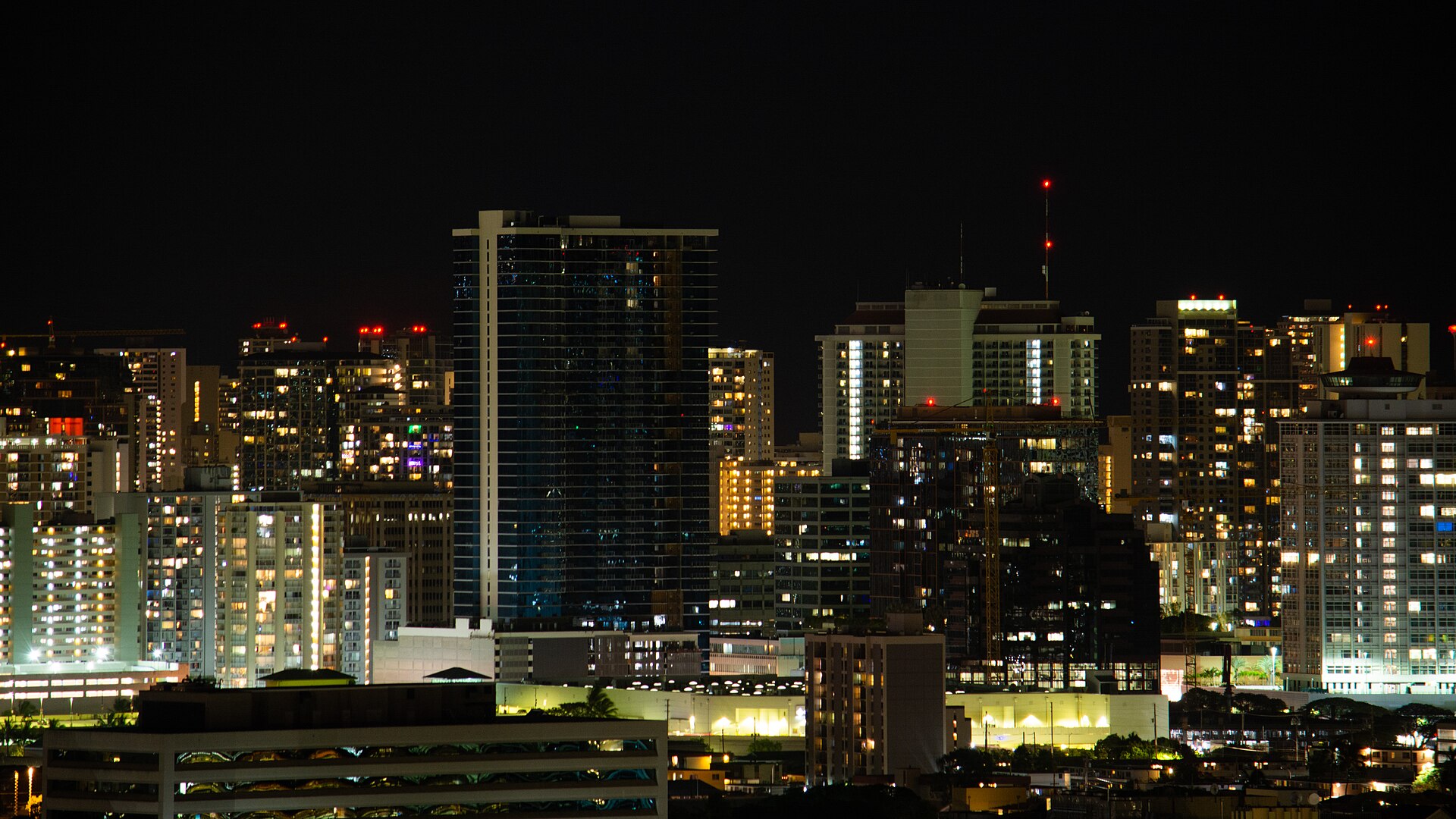 A cityscape image of Honolulu, Hawaii, at night time. The top third of the image depicts black night sky, with the bottom two thirds occupied by buildings of varying heights shapes and types. Most of the buildings are lit from inside, and the main pops of colour in the picture come from the red, white, yellow, orange, and blue lights.
