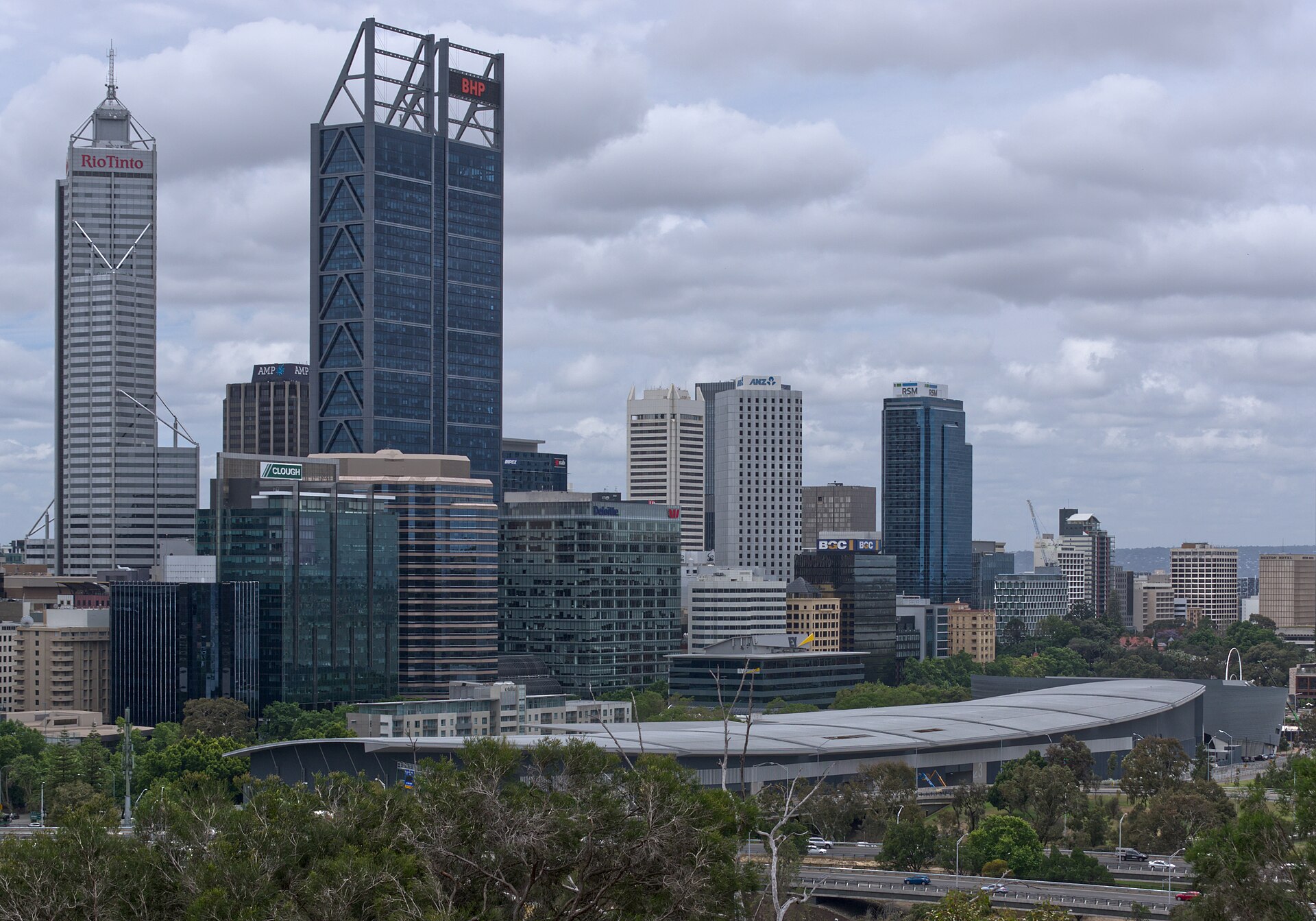 A cityscape of Perth. The foreground shows a row of the tops of trees, as though the image is taken above ground level short distance away. Large buildings can be seen on the left, and the buildings decrease in size as they get further into the distance. The picture is taken from an angle, closer to the left and looking out towards the horizon in the distance on the right. It is a cloudy day. 