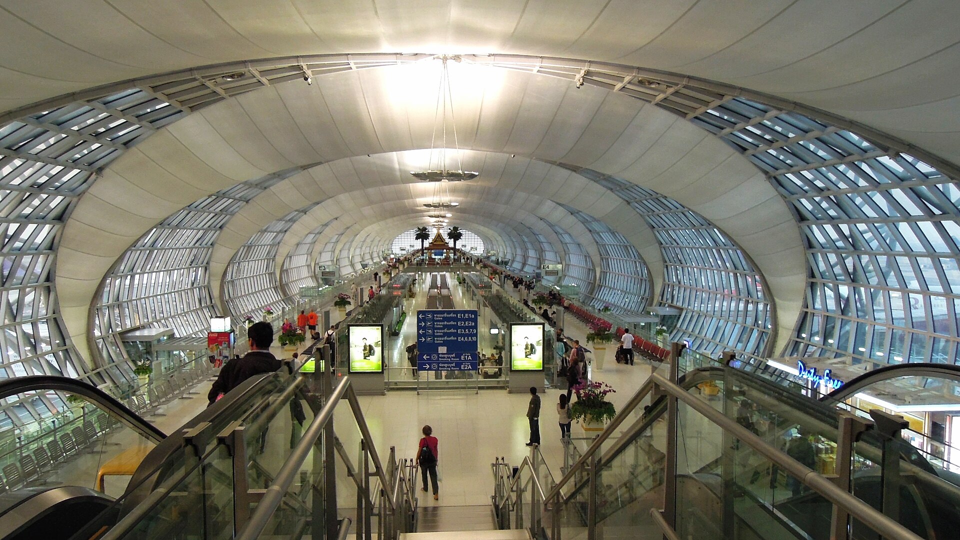 A wide corridor in Suvarnabhumi Airport. The image is taken from the top of the stairs at a far end of the corridor, giving a wide perspective. The walls are curved and diverge away from one another, creative the image of an oval shaped corridor with a flat floor. It is bright, and the walled are adorned with curved triangle windows that converge with one on the opposite side at th centre of the ceiling.
