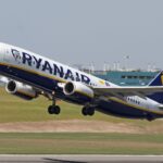 A Ryanair Boeing 737 taking off from an airport. The plane is mostly white with a navy blue underbelly and yellow stripe, as well as navy RYANAIR branding across the side of the fuselage and the company's logo on the navy blue tail. The nose is on the upper left of the image and the tail is lower centre right. The foreground shows tarmac surrounded by yellowing grass. The background shows a blueish grey sky.