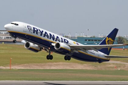 A Ryanair Boeing 737 taking off from an airport. The plane is mostly white with a navy blue underbelly and yellow stripe, as well as navy RYANAIR branding across the side of the fuselage and the company's logo on the navy blue tail. The nose is on the upper left of the image and the tail is lower centre right. The foreground shows tarmac surrounded by yellowing grass. The background shows a blueish grey sky.
