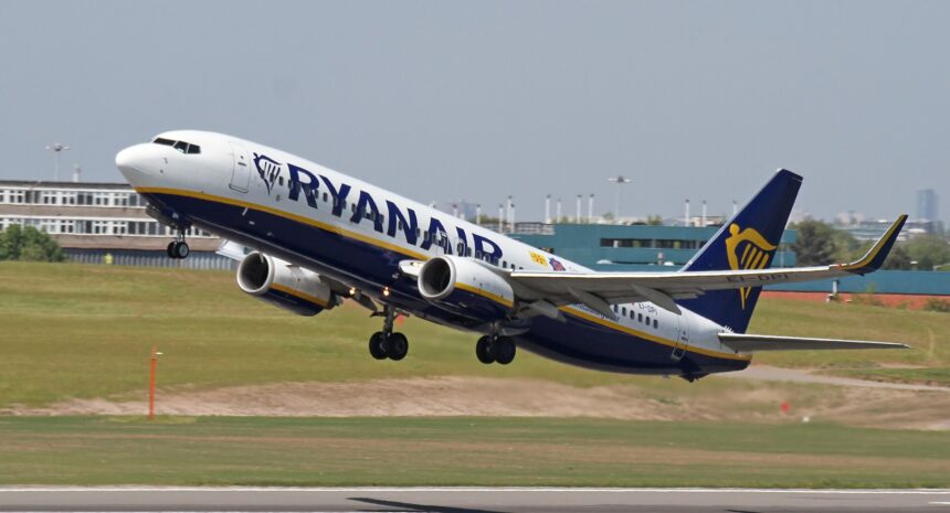 A Ryanair Boeing 737 taking off from an airport. The plane is mostly white with a navy blue underbelly and yellow stripe, as well as navy RYANAIR branding across the side of the fuselage and the company's logo on the navy blue tail. The nose is on the upper left of the image and the tail is lower centre right. The foreground shows tarmac surrounded by yellowing grass. The background shows a blueish grey sky.