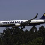 A Ryanair plane is pictured in flight. The aircrfat has the customary company branding across the side of the fuselage and tail in navy blue and yellow-gold. The jet's underbelly is also navy blue. The nose is facing left and the tail is on the right. In the background of the image is a clear blue sky.