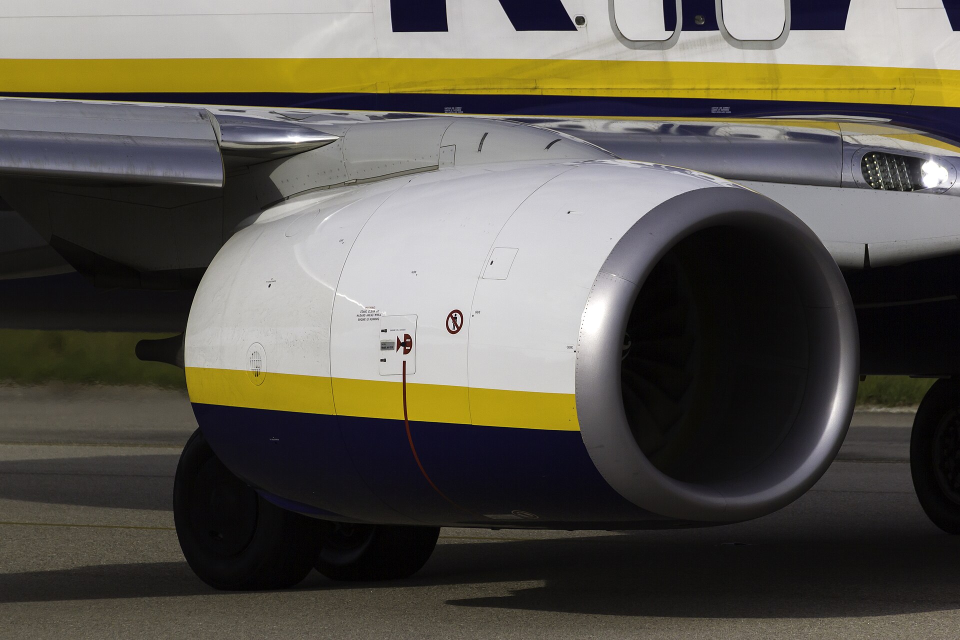 A Boeing 737 engine is pictured as the aircraft is stationed on airport tarmac. The engine is mostly white on the outside, with the bottom of it being navy blue, and a yellow strip separating the blue bottom from the white top. This colour branding is Ryanair, a British budget carrier. The engine is pictured from a left leaning angle, meaning the propellors are partly visible in the shadow of the centre. 