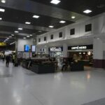 Terminal 2 of Manchester Airport. A row of shops can be seen lineing either wall of the terminal, with seating in the centre and walkways either side. The walls are predominently grey aside from the branding of the shops, and the scene is lit by fluorescent white overhead lights lined across the ceiling.