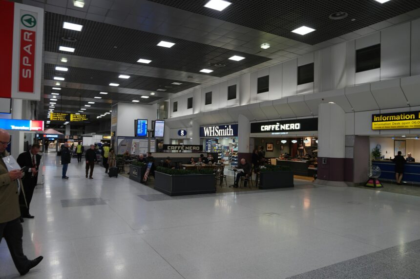 Terminal 2 of Manchester Airport. A row of shops can be seen lineing either wall of the terminal, with seating in the centre and walkways either side. The walls are predominently grey aside from the branding of the shops, and the scene is lit by fluorescent white overhead lights lined across the ceiling.