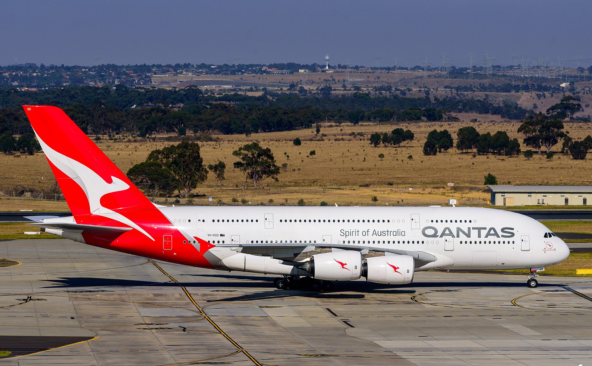 A Quantas plane on the ground at an airport. It is mostly white with large grey branding on the side, with red white and grey branding on the tail, which is on the left. The foreground is the grey concrete of an airport taxiway or runway. The background shows fields and a row of trees in front of a distant city. The top strip of the image is blue, and it appears to be a sunny day. 