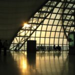 A large triangular window at Suvarnabhumi Airport in Thailand is the main focus of the image, stanning from the centre to the centre right. It appears to be sunset, outside, and a lovely golden hue is visible as the light pours in. There are squared lines across the triangle window, and in the centre left, the lower right corner of another of these windows can be seen. There are chairs underneath the window on the inside, where this picture is taken across the room from, and a cylinder which resembles a bin in near the centre of the image. All of the objects are silhouettes.