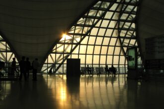 A large triangular window at Suvarnabhumi Airport in Thailand is the main focus of the image, stanning from the centre to the centre right. It appears to be sunset, outside, and a lovely golden hue is visible as the light pours in. There are squared lines across the triangle window, and in the centre left, the lower right corner of another of these windows can be seen. There are chairs underneath the window on the inside, where this picture is taken across the room from, and a cylinder which resembles a bin in near the centre of the image. All of the objects are silhouettes.
