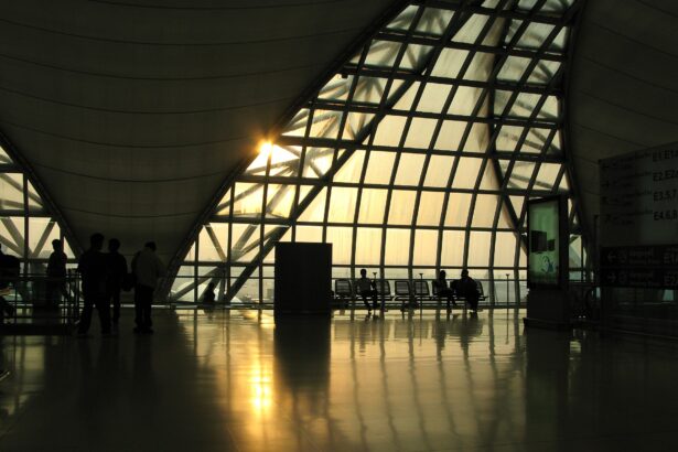 A large triangular window at Suvarnabhumi Airport in Thailand is the main focus of the image, stanning from the centre to the centre right. It appears to be sunset, outside, and a lovely golden hue is visible as the light pours in. There are squared lines across the triangle window, and in the centre left, the lower right corner of another of these windows can be seen. There are chairs underneath the window on the inside, where this picture is taken across the room from, and a cylinder which resembles a bin in near the centre of the image. All of the objects are silhouettes.