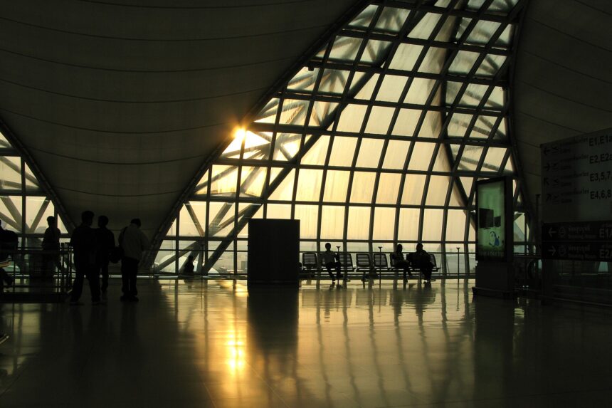 A large triangular window at Suvarnabhumi Airport in Thailand is the main focus of the image, stanning from the centre to the centre right. It appears to be sunset, outside, and a lovely golden hue is visible as the light pours in. There are squared lines across the triangle window, and in the centre left, the lower right corner of another of these windows can be seen. There are chairs underneath the window on the inside, where this picture is taken across the room from, and a cylinder which resembles a bin in near the centre of the image. All of the objects are silhouettes.