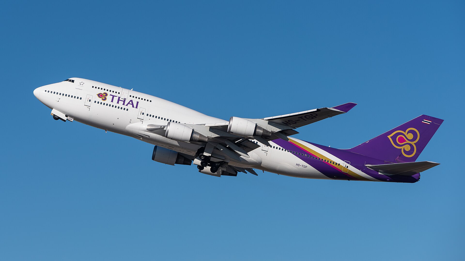 A Thai Airways International Boeing 747 aircraft is ascending. It is mostly white with purple worded branding along the side, and a completely purple, gold and red tail to accompany this. The nose is upper left centre and the tail is lower right centre. The background is cloudless blue sky.