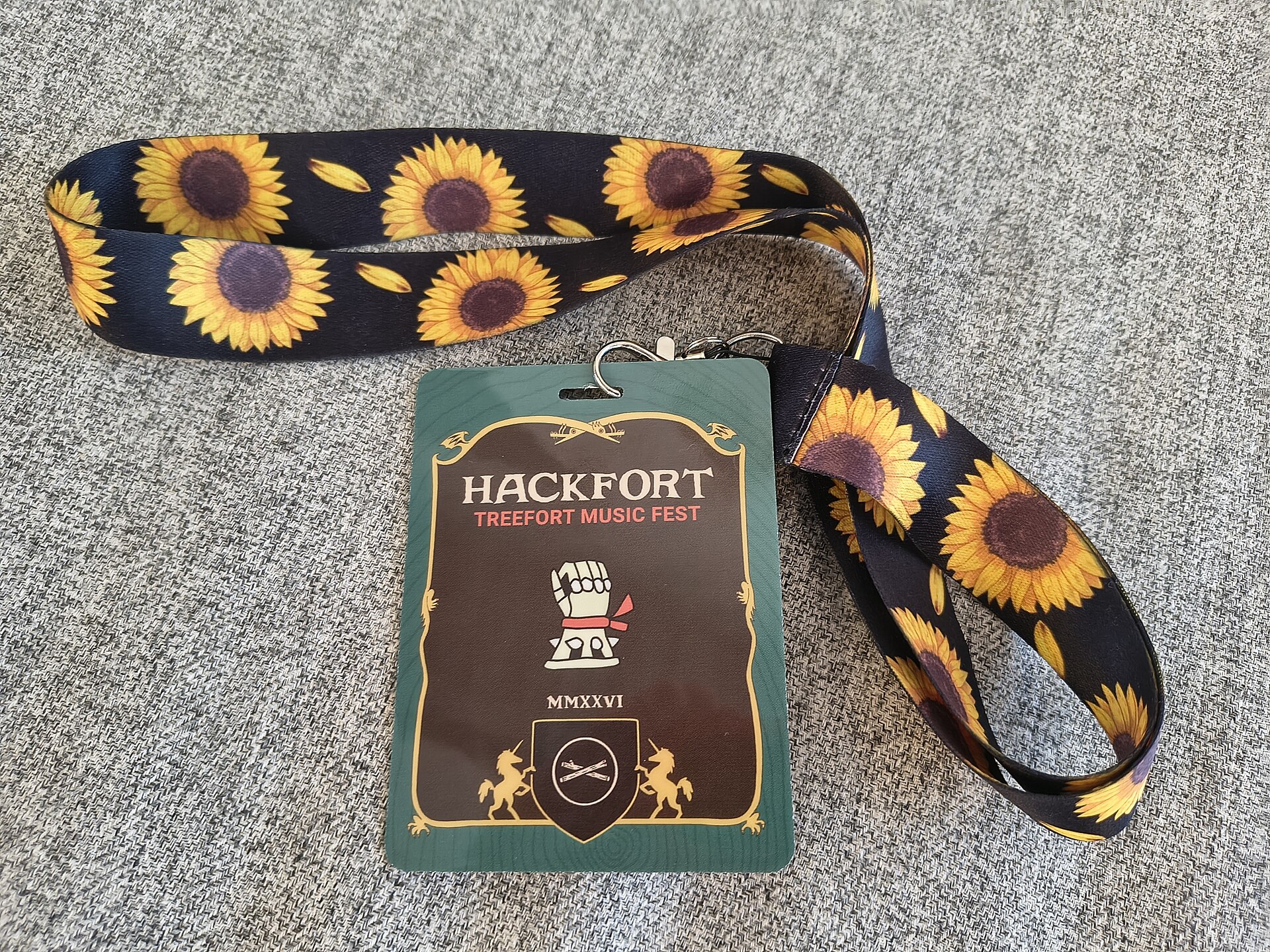 An image of a pass to a music festival attached to a black lanyard with sunflowers on it. The pass is green and brown. The background is a plain beige fabric which is most likely a carpet. 