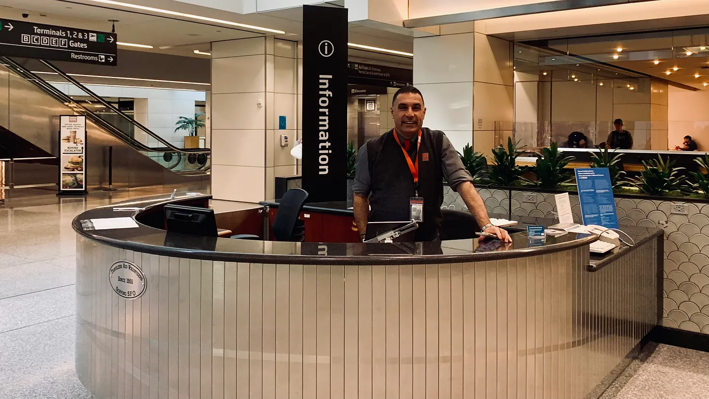 Information help desk as the staff member stands behind the desk with a smile on his face, with an escalator and airport shops that can be seen in the background