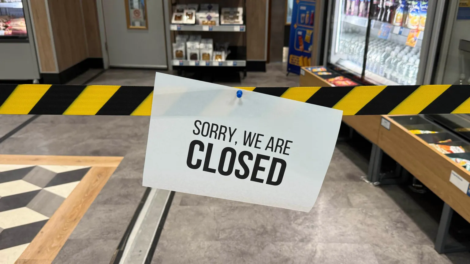 A paper sign saying "sorry, we are closed" hangs at the entrance to a sandwich shop on black and yellow striped warning tape at Gatwick Airport.