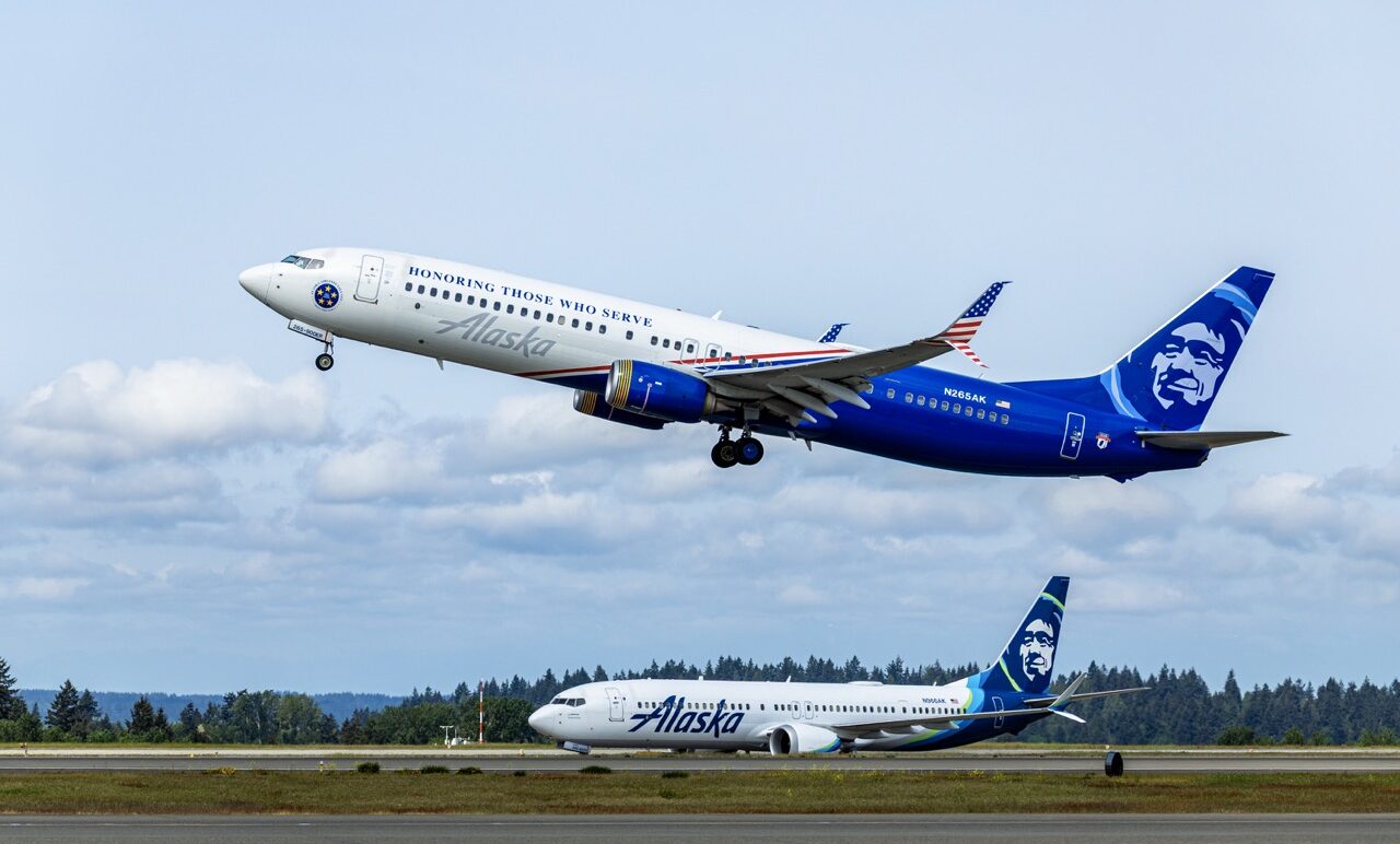 Alaska Airlines aircraft taking off, with another underneath it. White and blue livery with American flag stripes on the fuselage middle. "Honoring those who serve" written in blue.