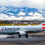 An American Airlines aircraft parked in front of very snowy mountains. The sky is cloudy, and, behind the airplane and in front of the mountains, the grass is a mixture of green and brown.