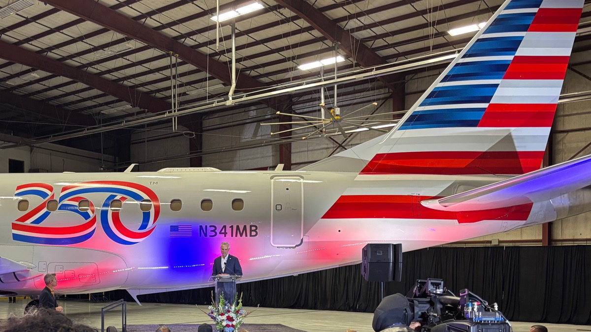 The tail of a white plane which has the American flag design. 250 written in blue, red and white on the fuselage. A man stood in front of the plane, talking behind a podium. 