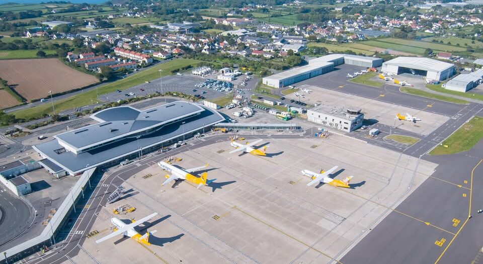 An aerial shot of Guernsey Airport. Yellow and white aircraft seen on the apron. Houses and buildings seen in the background toward the top.