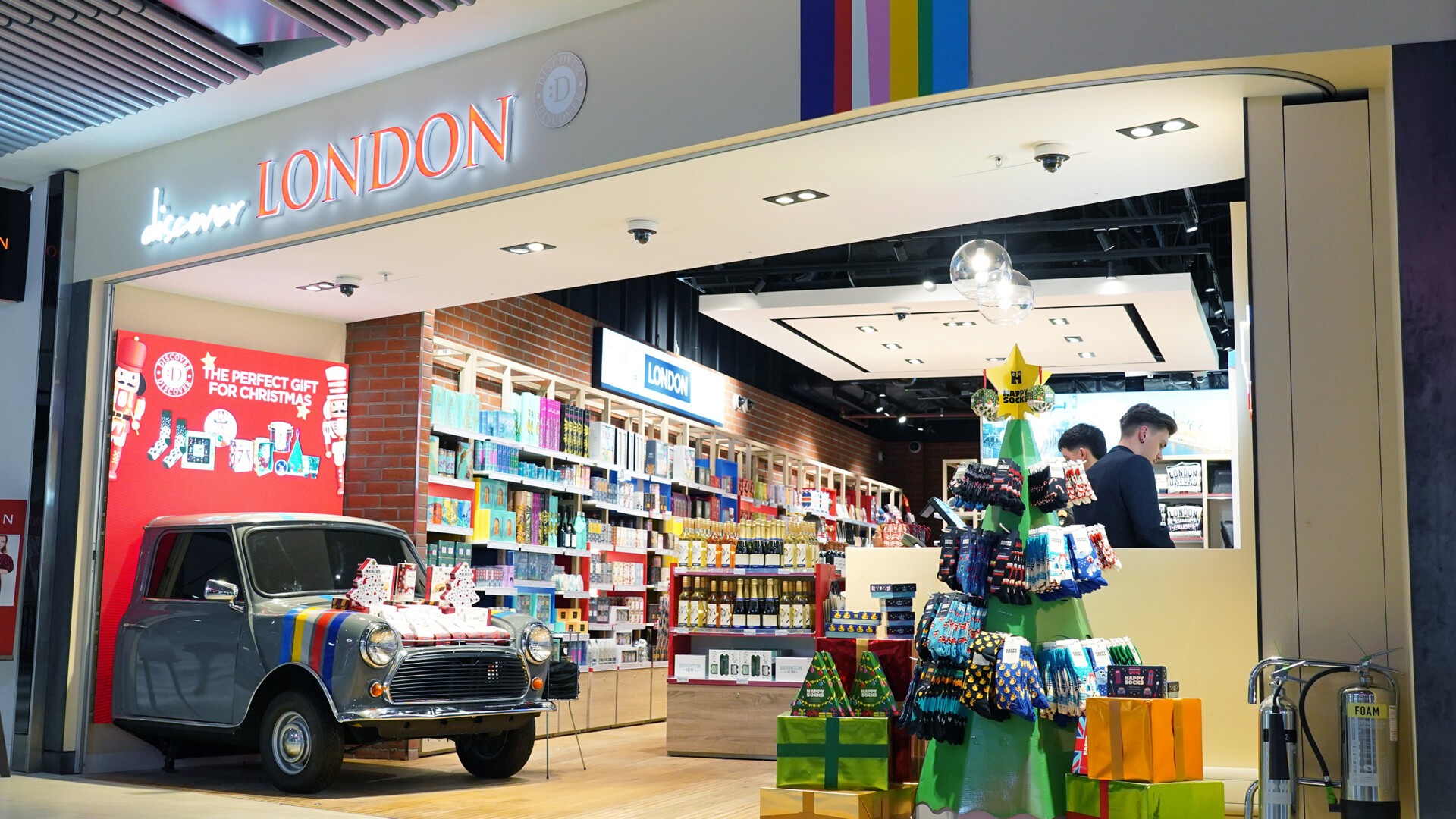 Image shows 'discover London' shop front at London Gatwick Airport (LGW) south terminal with half a Mini Cooper car, stocked shelves and shoppers