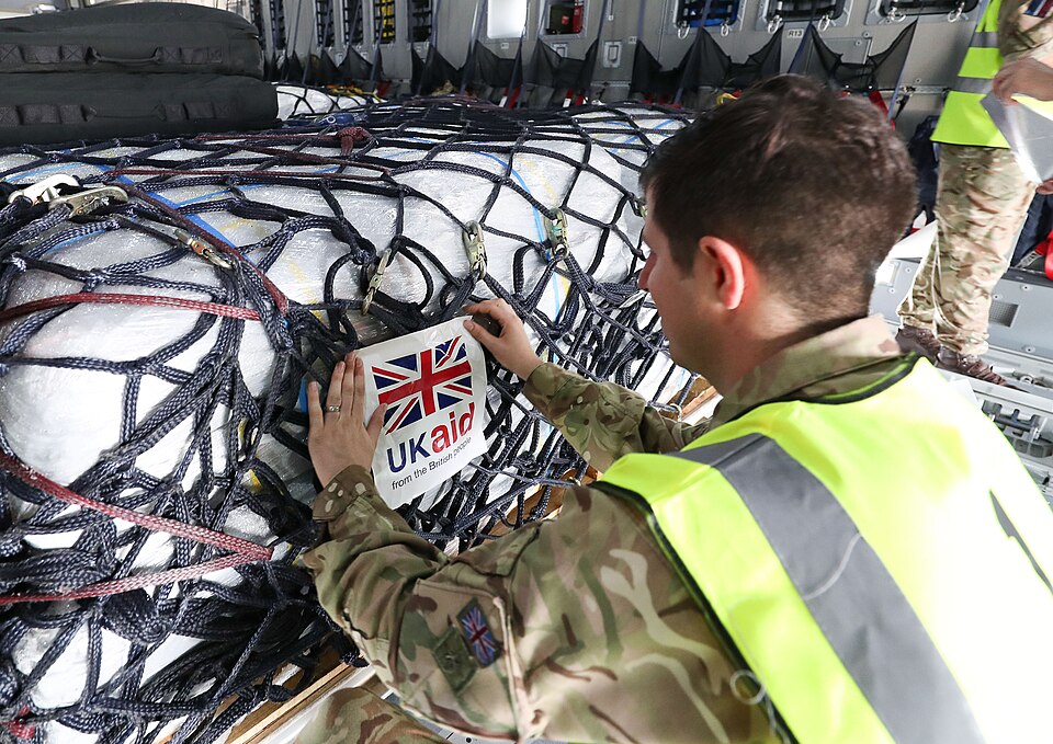 A British military service member wearing camouflage fatigues and a high-visibility vest attaches a "UK aid from the British people" label to large cargo pallets secured with netting inside the hold of a transport aircraft.