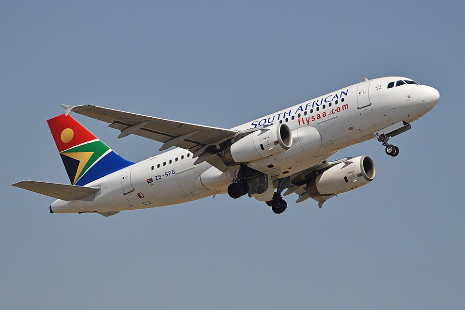 A detailed, low-angle view of a twin-engine South African Airways Airbus A319-131 passenger jet climbing in mid-air. The aircraft features a predominantly white fuselage with "SOUTH AFRICAN" written in blue text.