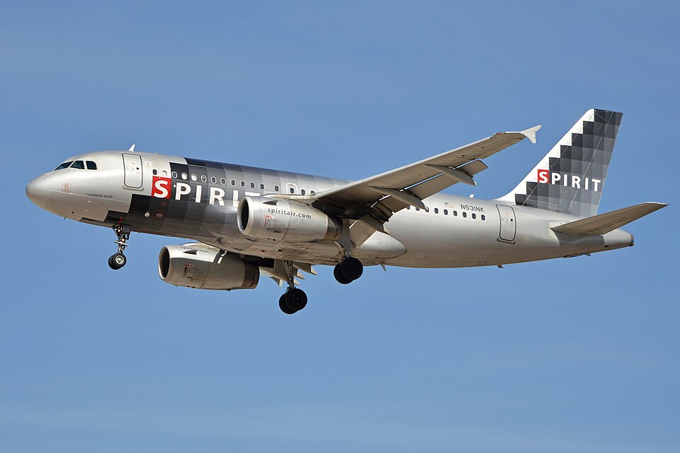 A Spirit Airlines aircraft flying under a blue sky. its livery is a mix of dark and light grey with tiny red accents