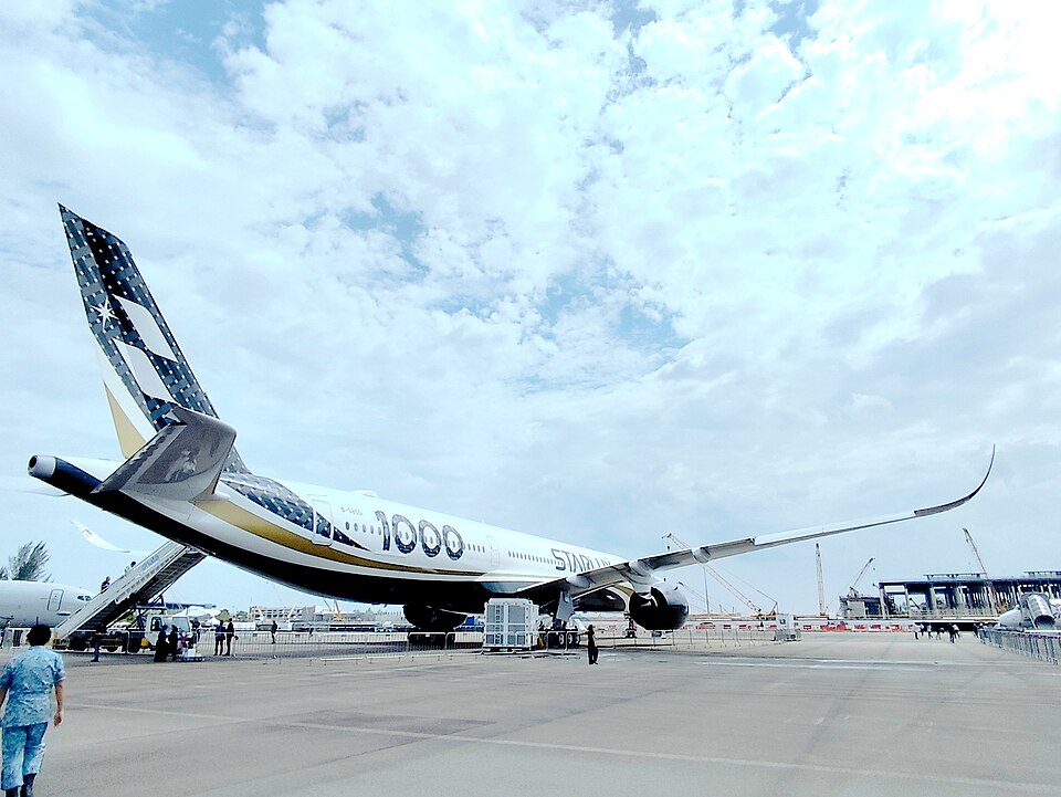 A Starlux Airlines Airbus A350-1000 parked on a tarmac, viewed from a low, wide-angle rear-left perspective. The aircraft displays its distinctive livery, featuring a white fuselage with large "1000" branding, and a dark tail section adorned with a stylized compass star logo and a sweeping gold band. The long right wing with its curved winglet stretches out prominently. Mobile passenger stairs are attached to the rear door, and metal crowd barriers surround the aircraft, suggesting a public display or airshow setting, with airport construction and cranes visible in the background under a bright, heavily clouded sky.
