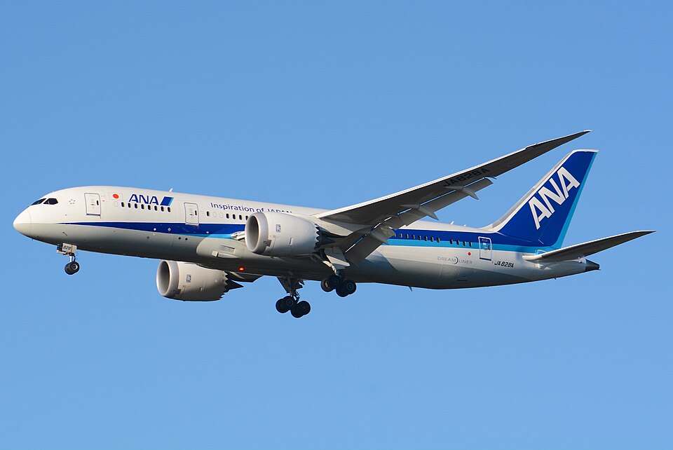 An ANA (All Nippon Airways) Boeing 787 Dreamliner (registration JA828A) on approach with landing gear extended, featuring the airline's white and blue livery with "Inspiration of Japan" slogan, set against a clear blue sky.