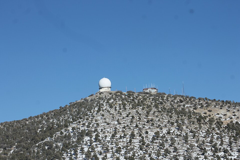 A large white geodesic radome and several communication towers situated on a rugged, snow-dusted mountain peak against a clear blue sky.