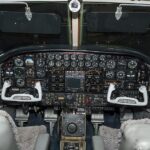 A detailed interior view of a vintage aircraft cockpit. The complex, dark instrument panel is densely packed with rows of traditional circular analog gauges, switches, and a central radio/navigation stack. Two distinct, white, U-shaped control yokes are positioned in front of pilot and co-pilot seats featuring grey sheepskin covers. The throttle quadrant and various control wheels sit on the center pedestal between the seats, and two dark sun visors are pulled down over the windshield against a dark background outside