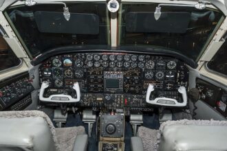A detailed interior view of a vintage aircraft cockpit. The complex, dark instrument panel is densely packed with rows of traditional circular analog gauges, switches, and a central radio/navigation stack. Two distinct, white, U-shaped control yokes are positioned in front of pilot and co-pilot seats featuring grey sheepskin covers. The throttle quadrant and various control wheels sit on the center pedestal between the seats, and two dark sun visors are pulled down over the windshield against a dark background outside