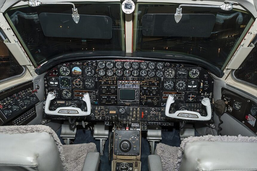 A detailed interior view of a vintage aircraft cockpit. The complex, dark instrument panel is densely packed with rows of traditional circular analog gauges, switches, and a central radio/navigation stack. Two distinct, white, U-shaped control yokes are positioned in front of pilot and co-pilot seats featuring grey sheepskin covers. The throttle quadrant and various control wheels sit on the center pedestal between the seats, and two dark sun visors are pulled down over the windshield against a dark background outside