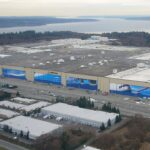An aerial view of a massive Boeing manufacturing facility, characterised by its sprawling flat roof and enormous hangar doors adorned with large, blue aviation-themed murals and the Boeing logo. The massive complex is situated among smaller white-roofed industrial buildings, parking lots, and roads in the foreground, with a landscape of forested areas, a wide body of water, and distant landmasses visible in the background under a hazy sky.